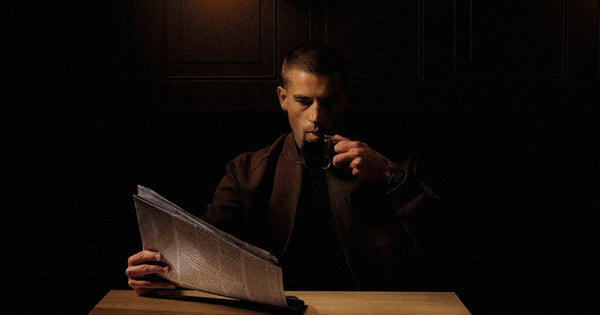 A man reading a newspaper late at night under a single light, drinking an espresso