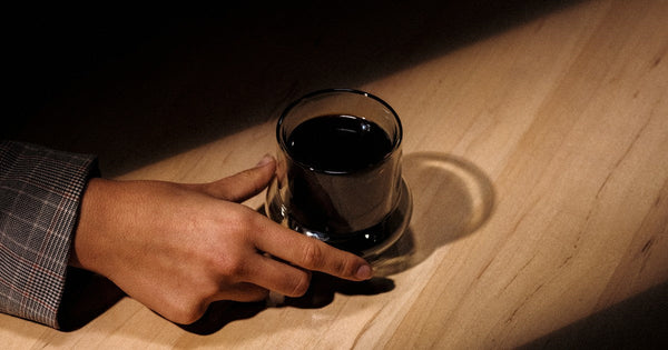 A hand cradling a glass of black coffee resting in a pool of warm light on a wooden table